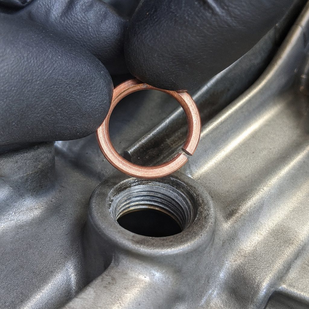 gros plan sur un joint de vidange en cuivre fendu tenu entre deux doigts gantés, la fente bien visible, au-dessus d'un carter moteur en aluminium